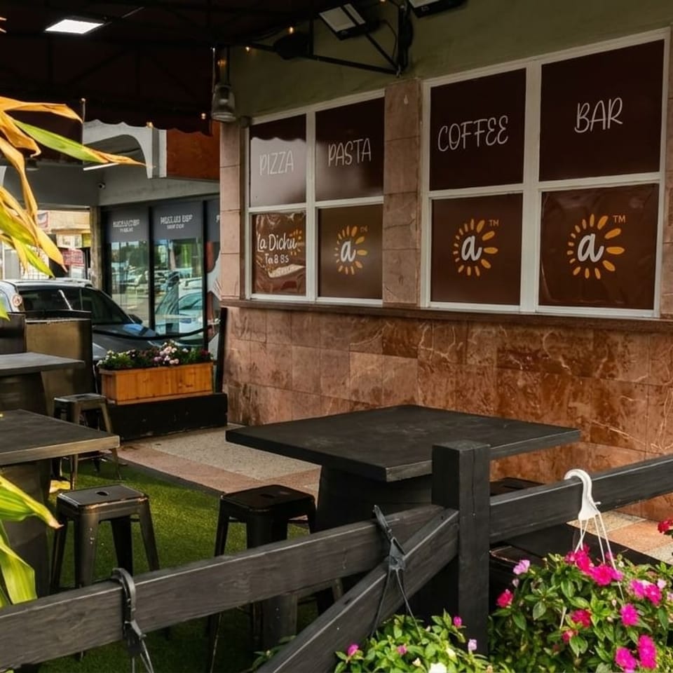Outdoor patio of La Dicha restaurant with tables, stools, plants, and window signs for pizza, coffee, and bar.