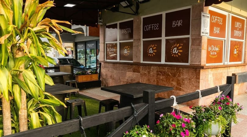 Outdoor patio of La Dicha restaurant with tables, stools, plants, and window signs for pizza, coffee, and bar.