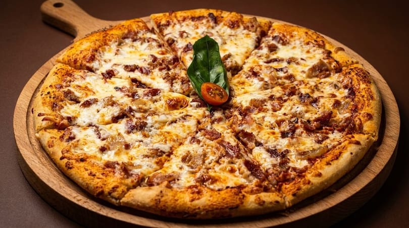 A freshly baked pizza with melted cheese, bacon, and onions, garnished with basil and cherry tomato.