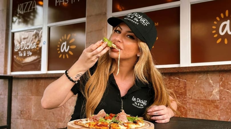 Woman in a La Dicha hat and shirt eating a slice of pizza with stringy cheese.
