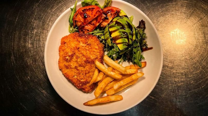 Crispy fried chicken schnitzel with golden fries, avocado, grilled tomatoes, and a green salad.