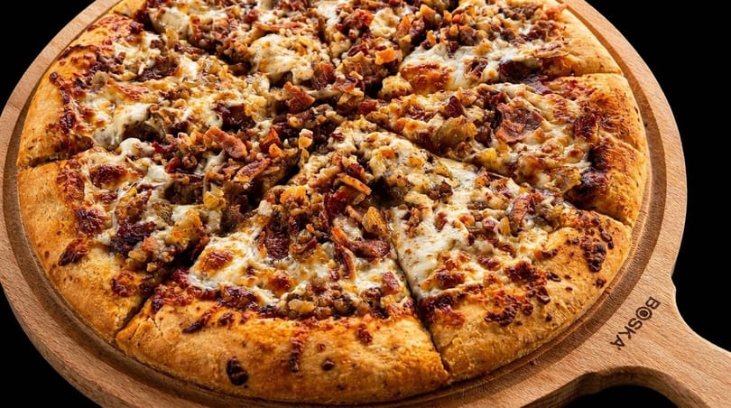 A sliced pizza topped with melted cheese, bacon, and onions on a wooden serving board.