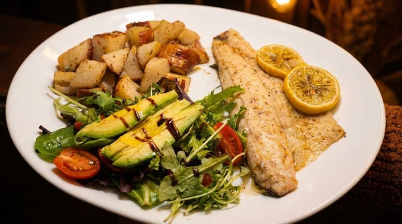 Grilled fish with lemon slices, roasted potatoes, and a fresh salad with avocado and balsamic glaze.