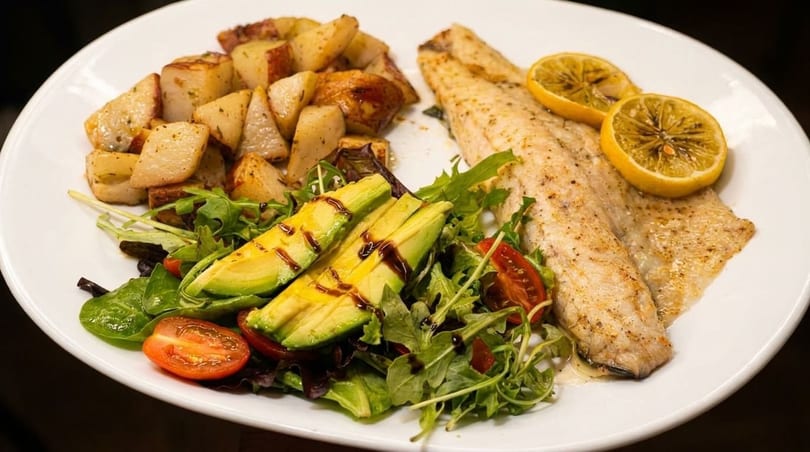 White plate with baked fish, roasted potatoes, avocado, and a fresh green salad.