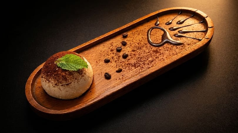 Tiramisu dessert with cocoa powder, mint, coffee beans, and chocolate drizzle on a wooden tray.