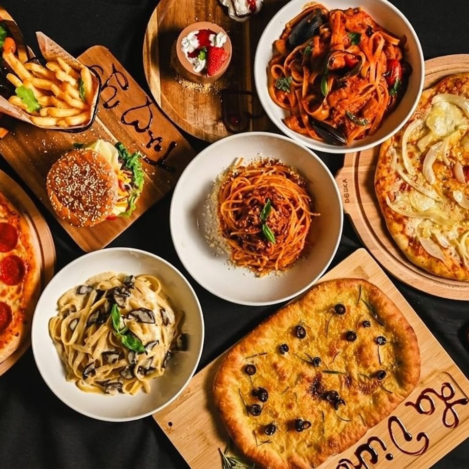 Overhead view of a feast of pizzas, pastas, salads, a burger, fries, and dessert on a dark surface.