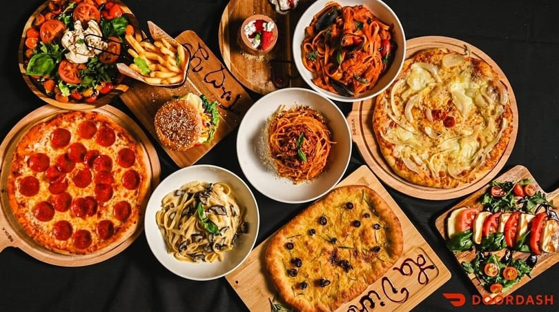 Overhead view of a feast of pizzas, pastas, salads, a burger, fries, and dessert on a dark surface.